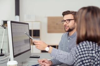 Proficient young male employee with eyeglasses and checkered shirt, explaining a business analysis displayed on the monitor of a desktop PC to his female colleague, in the interior of a modern office.jpeg