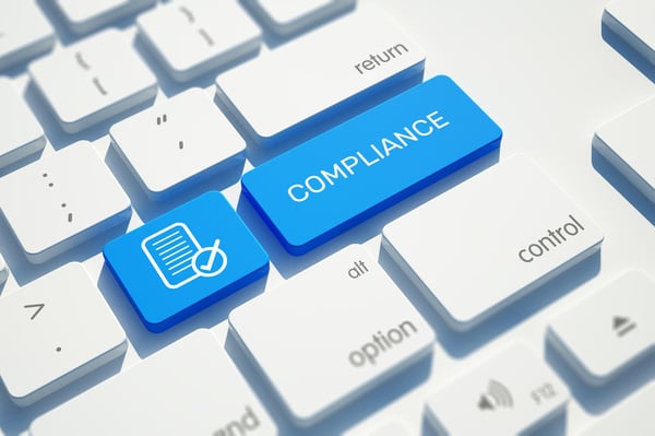 A white computer keyboard features a prominent blue key labeled "Compliance," accompanied by a document verification icon